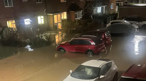 A picture that appears to have been taken from a first floor window, looking down on flooding in roads and into homes. There appears to be water about half way up cars' wheels 