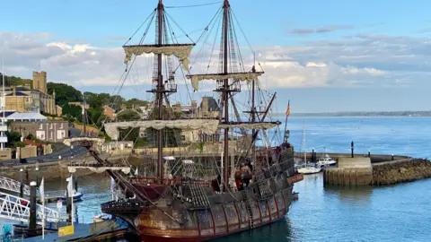 A large 17th Century Spanish galleon replica ship is seen docked at Cowes, with buildings and the harbour in the background under a mostly blue sky. 