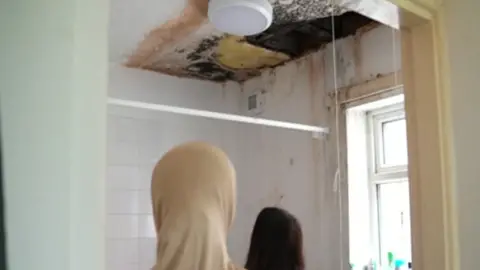 Two women look up at a black mouldy ceiling above a bathroom. The backs of their heads are pictured through a doorway. One wall is tiled in white