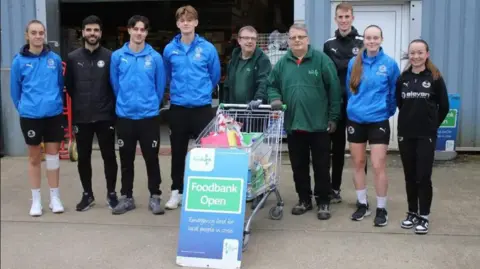 Four football students wearing shorts and blue tops stand with three adults wearing black top and tracksuit bottoms, alongside two adults in green fleece jackets. A shopping trolley full of food is in front of them all. They are outside a warehouse.