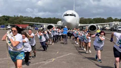 A group of people split in two are pulling a plane by two ropes on a slightly overcast day. A warden wearing a blue top and a hat can be seen in the middle of the two groups, in front of the airplane. Lorries and another plane can be seen in the distance.