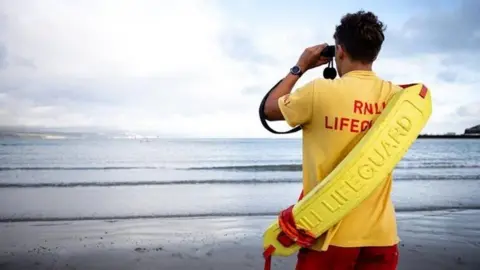 RNLI Lifeguard with red shorts and yellow top with yellow rescue tube across his back looking out to the sea off Weymouth with binoculars