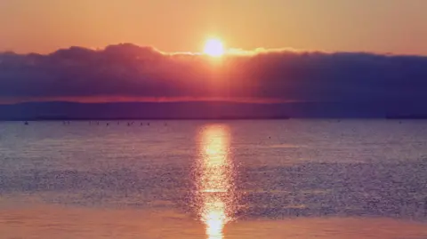 Lesley FRIDAY - A vivid sunrise over Weymouth shows an orange sun reflected in the sea off the Dorset coast causing the water and sky to glow orange. In the distance a low bank of cloud hovers over the dark cliff