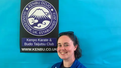 Sinéad Byrne smiling in front of blue wall and sign saying Kenpo karate & Budo Taijutsu club. 