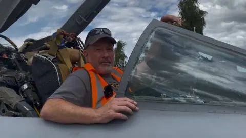 A man, with grey hair and a beard, sits in the cockpit of the Lightning jet. He is wearing a hi-vis jacket and a baseball cap with sunglasses. There is flying equipment behind him.