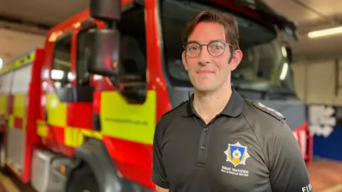 A man wearing a top with a West Yorkshire Fire and Rescue logo stands in front of a fire engine. He has dark hair and glasses.