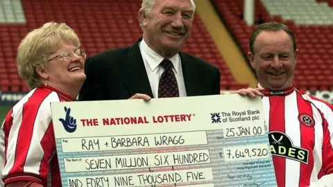 PA A couple dressed in red and white Sheffield United shirts stand either side of a suited man holding a giant cheque. The cheque reads: "Ray + Barbara Wragg; Seven million, six hundred and forty nine thousand' and is dated 25 January 2000.