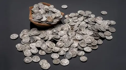 A large number of silver Roman coins in a wooden bowl and scattered around