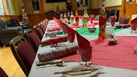 Glenn Johnson A table set for a community Christmas Day dinner in Ardess Parish Church hall in Kesh in December 2023.  The long table is laid with white, green and red table cloths, plates, glasses, cutlery, crackers and bottles of grape juice.