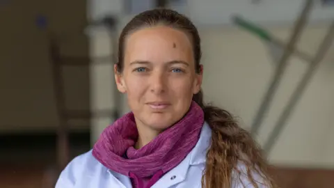 States of Guernsey Alex Tielles standing in an area of Guernsey Dairy while wearing a white laboratory coat and a pink coloured fleece underneath. Her brown hair is in a ponytail which is draped over her left shoulder.