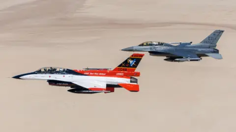 Two US Air Force F-16 combat jets fly side by side over the desert, one white and red, the other combat grey