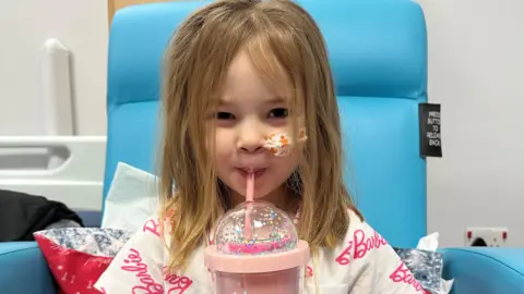 A girl with blonde hair and a white top with the word Barbie on it in pink lettering sitting in a blue chair and drinking something out of a pink cup with a straw