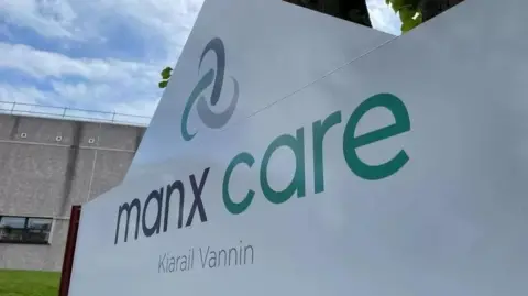 A white sign outside a hospital building that reads Manx Care in black and green font, with the Celtic knot logo above it. 

