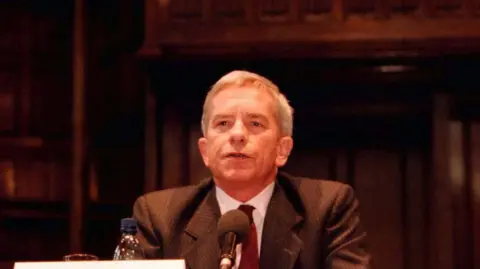 Press Association Lord Saville speaking into a mic at a hearing.  He is wearing a striped blazer, white shirt and a red tie.