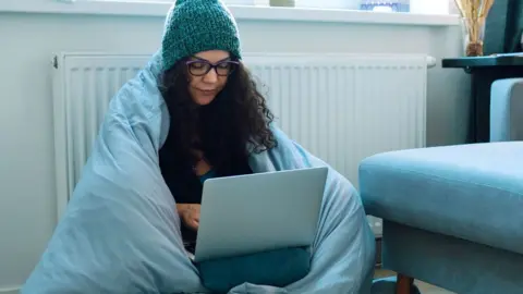 Women in a woolly hat and wrapped in a duvet sits next to a radiotor