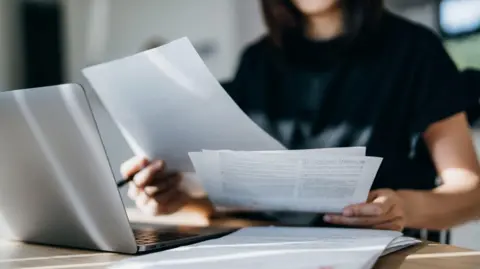 A woman sits holding paperwork in front of a laptop.