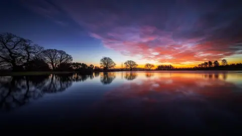 Hang Ross There is a large expanse of water with trees lining its distant shore. The trees are silhouetted against the sky, which is a dark blue colour but also bright orange on the horizon and peppered with pink clouds.