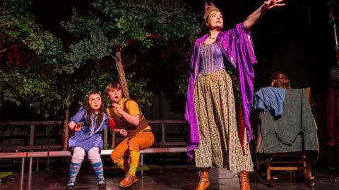 Bright Umbrella Theatre Company Three actors perform a scene on stage in East Belfast. In the foreground is a woman dressed as as witch in a paisley-patterned skirt, purple top and purple cloak and gold crown. Behind her two children cower - a girl with dark brown hair in a purple dress and purple tights and a boy with short brown hair in yellow socks, orange trousers, a yellow shirt and brown waistcoat