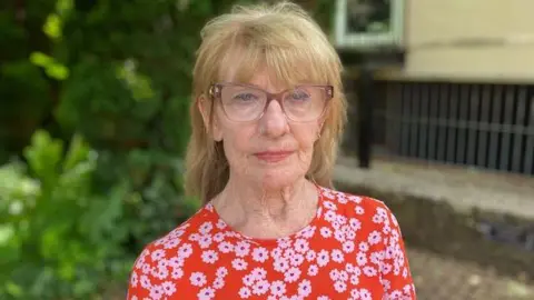  Jennifer Hill, who has blonde hair and large clear/purple framed glasses. She is wearing a red top with pink flowers on it.