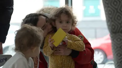 Brooke and her two-year-old daughter Ivy. Ivy holds a yellow toy and has brown curls, she looks into the camera as her mum Brooke snuggles her shoulder.