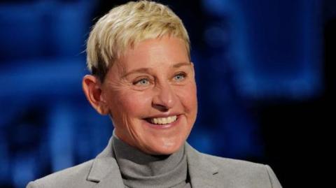 Ellen DeGeneres smiling, wearing a grey turtleneck top and grey jacket