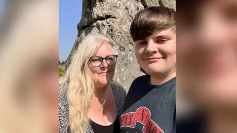 PA Dylan with his mum stood in front of a large rock in the countryside smiling.
Dylan is wearing a black t-shirt and his mum with wearing a black top and glasses.