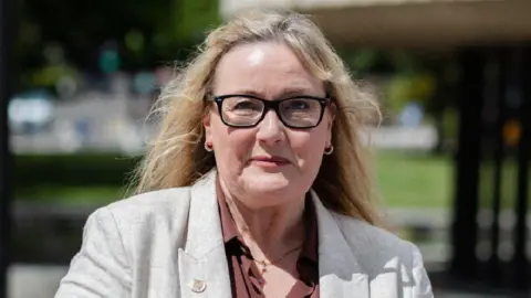 Karen Kilgour, who has long blonde hair and is wearing black glasses, a grey linen blazer and brown shirt, smiles at the camera.