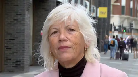 Melanie Leahy standing on the street. She has white hair and is wearing a baby pink blazer and black top.
