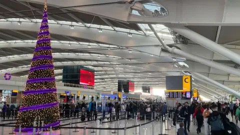 People with luggage walk in a Heathrow Airport terminal. A Christmas tree has purple decorations 