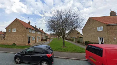 Google street view of the road facing Ormesby Walk which is a pathway with grass in the middle. There are several terraced houses along the path and two cars parked out front. 