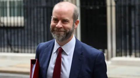 Reuters Jonathan Reynolds walking up Downing Street wearing a suit and tie and carrying a red folder.  