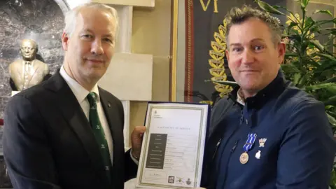 Gideon Amos Office two men looking at camera holding a certificate with a statue and a plant in the background