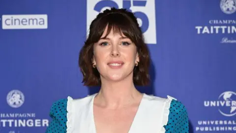 Alexandra Roach smiles at the camera wearing a white and blue dress, standing in front of a blue background. 