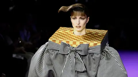 Model on the catwalk wears a black and white stripe top with oversized bow and a neck design resembling a chess board