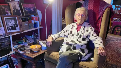 Glenys Cour sitting in a mustard armchair with a pink, red and purple blanket behind her. She has white short hair and is wearing pink glasses, pink earrings and a white blouse with black irregular spots. Besides her is shelving full of family photographs.