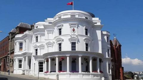 The outside of the white Tynwald building, known as the wedding cake building as it has several tiers. A red and white Manx flag flies on top of it with blue sky as a backdrop.
