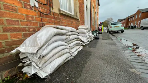 Ben Schofield/BBC Piles of white sandbags leaning against the fronts of terraced houses in Newport Pagnell. The image is taken looking along the pavement, which disappears into the distance. To the right of the image is the road, with some cars parked up to the sides. The houses are red brick, Victorian-looking terraces.