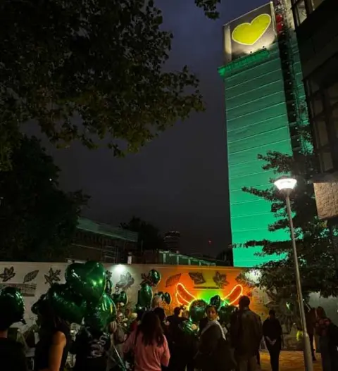 BBC An image of the Grenfell Tower lit up in green against the backdrop of a night sky in London on Wednesday night, with people gathered at the base