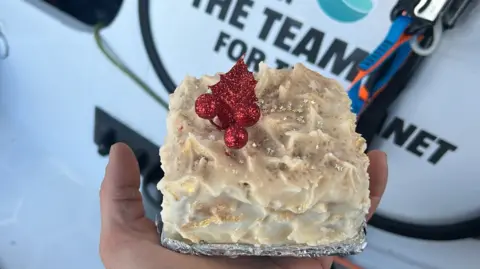 Sam Goodchild Christmas cake being held in one hand with a red decoration on top of the icing with some words in the background.