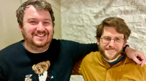 John (left) with his arm over Jon's (right) shoulder smiling at the camera 