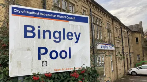 Close-up shot of a white street sign with Bingley Pool in large, blue lettering and a yellow Yorkshire stone building in the background.   