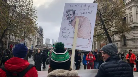 Protestors carry banners including one with a cartoon image of Prime Minister Keir Starmer's head with a rat's body. The words on the sign read 'Starmer fled Welsh farmers "like a rat".  Will he face us today?'