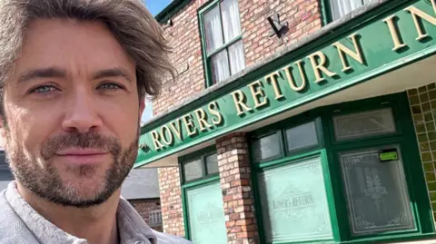 Emrhys Coopers stands in front of Coronation Street's Rovers Return Inn. The pub has a red brick exterior with a green facade. The letters 'Rovers Return Inn' are written in gold.