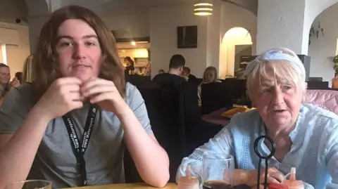 A young man with long brown hair smiles at the camera at a restaurant, sitting next to an elderly lady