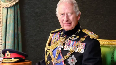 PA King Charles III sitting in the Grand Corridor at Windsor Castle, wearing his Field Marshal No1 Full Ceremonial Frock Coat with medals, sword and decorations. His Field Marshal cap, white gloves and baton are placed on the table beside him.