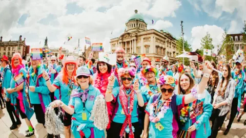 Volunteers in blue shirts and brightly coloured headgear wave flags in front of Hull City Hall