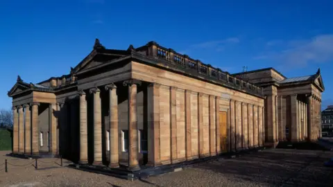 A general view of the National Gallery of Scotland in Edinburgh. It is a sunny day with blue sky and a few grey-white clouds in the sky. The building itself is made from light stone and has two sets of four columns on either side.