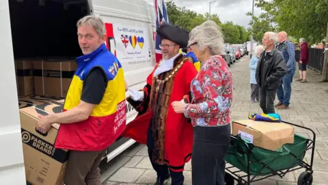 A load of people loading up a van with a mayor in official clothing watching