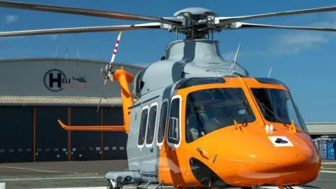 A yellow and grey helicopter in front of a HeliOperations base.