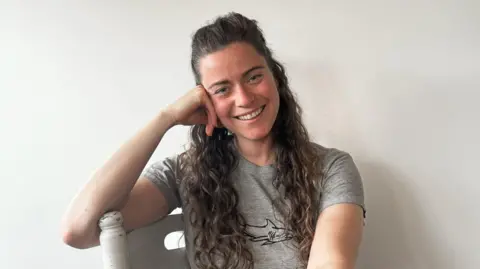 Charlie Gill Charlie Gill rests her head on her hand while leaning on a white painted chair. She wears a grey T-shirt with a shark printed on it and smiles at the camera. She has long curly brown hair which she wears half-up-half-down